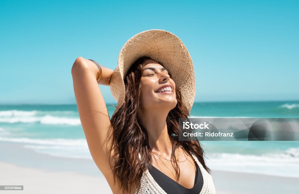 Stylish casual woman enjoying sun at tropical beach Beautiful girl with straw hat enjoying sunbath at beach. Close up face of young tanned woman with closed eyes enjoying breeze at seaside. Carefree latin woman smiling with ocean in background. Women Stock Photo