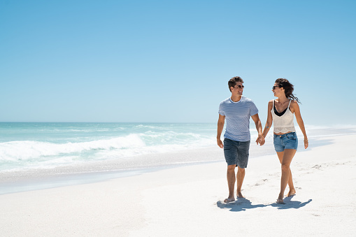 Happy young couple holding hands and walking barefoot on beach with copy space. Handsome man and beautiful woman in casual wearing sunglasses at seaside. Cheerful smiling couple relaxing on the sea shore of a white sand.