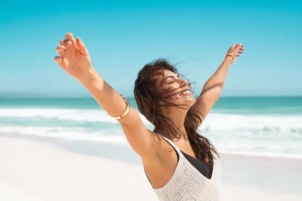 Portrait of healthy young woman standing on the beach with outstretched arms and feeling the breeze. Happy young woman feeling fresh and relaxing at ocean. Latin tanned woman with closedd eyes feeling good.