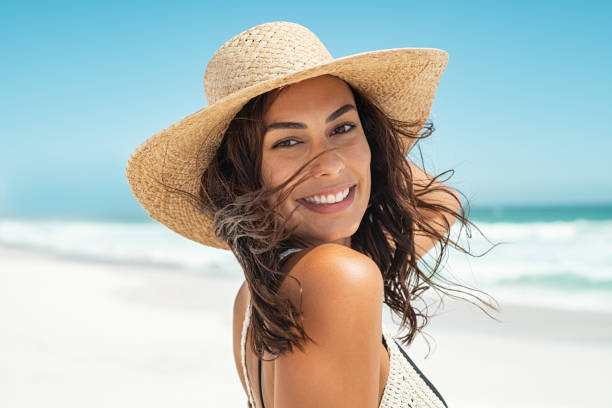Carefree stylish woman enjoying summer Portrait of beautiful smiling young woman wearing straw hat at beach with sea in background. Beauty fashion girl looking at camera at seaside with big grin. Carefree tanned woman walking on sand and laughing. beautiful woman summer stock pictures, royalty-free photos & images