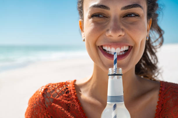Young happy woman drinking soft drink on beach Close up face of young woman drinking fresh sparkling water from a glass bottle at beach. Portrait of beautiful carefree girl drinking soft drink with straw during summer. Smiling girl staying hydrated during summer with lemonade. soda bottle photos stock pictures, royalty-free photos & images