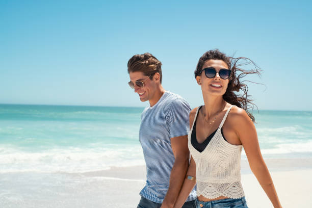 Young couple enjoy summer vacation Beautiful woman with man wearing sunglasses walking on beach.. Young couple enjoying honeymoon after marriage at sea. Happy casual couple holding hands and walking at the beach with copy space. young couple stock pictures, royalty-free photos & images