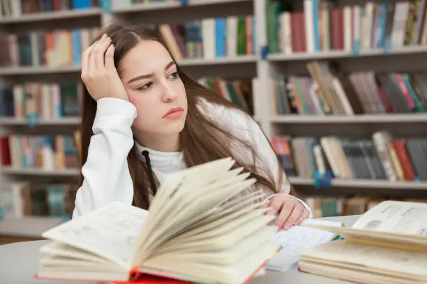 Teenage girl looking bored while studying at the library. Tired young girl disliking learning. Sadness concept