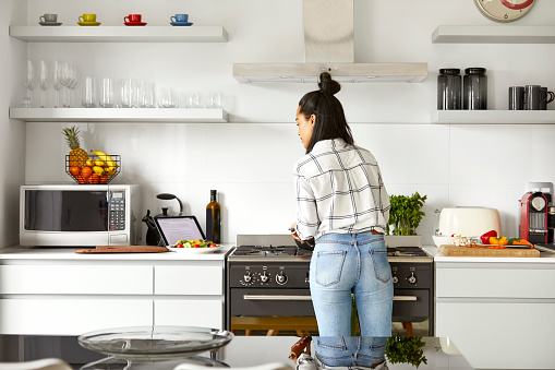 Rear view of woman looking at digital tablet while cooking food. Female is learning new recipe through internet. She is standing in kitchen at home.