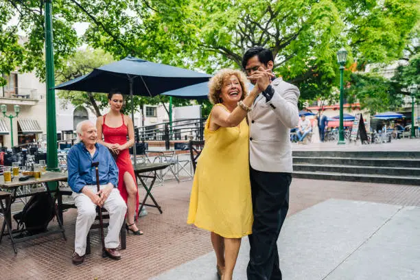 Smiling senior woman dancing the tango with skilled young man while her husband and young female dancer watch.