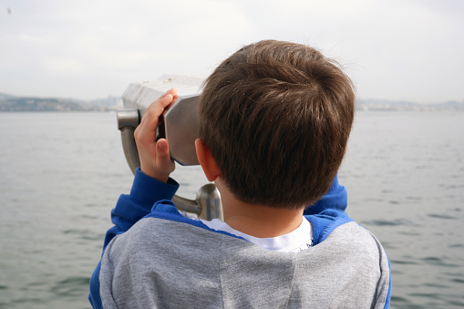 A curious, nine year old caucasian boy looking through coin operated binoculars to a seascape in spring.