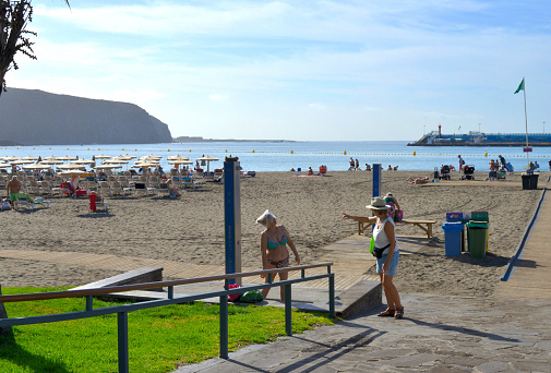 Los Cristianos, Tenerife/ Spain -  February 07, 2020: senior lady Rain cap on head in bikini taking a shower on the beach while talking to another woman showing something with her finger. Swimming area demarcation. Lounge chairs on beach. Children carriage and mothers.