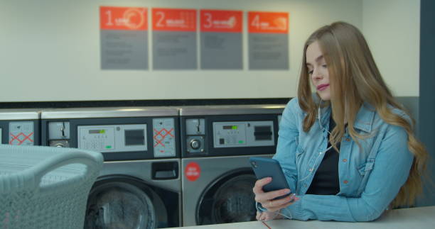 istock Woman using smartphone while washing her laundry at laundromat. 1211347221