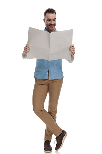 Happy casual man reading newspaper and smiling while wearing blue shirt, standing on white studio background