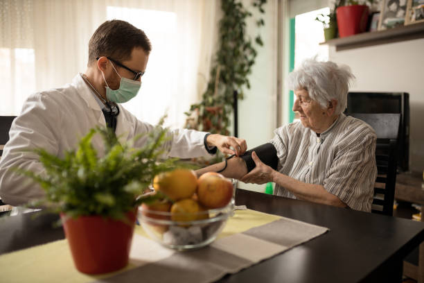Young doctor adjusting blood pressure gauge on senior woman's hand during house call visit Young serious Caucasian doctor wearing face mask adjusting blood pressure gauge on senior gray-haired woman's hand during house call medical check-up hypertensive photos stock pictures, royalty-free photos & images