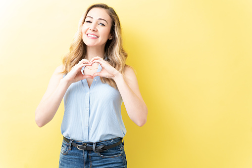 Cheerful adult woman making heart shape with hands while standing against yellow background