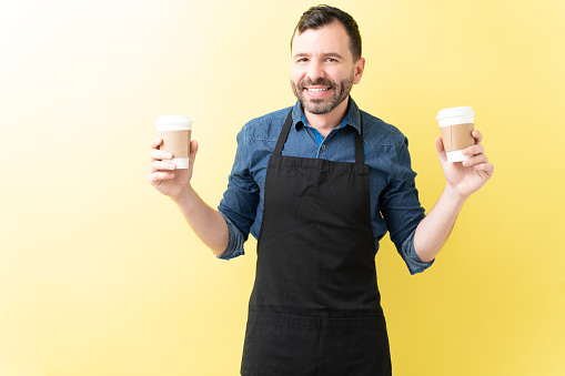 Smiling Hispanic male barista holding coffee paper cups over yellow background