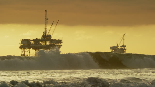 Slow Motion Shot of Waves Crashing onto the Shores of Huntington Beach in Southern California with Several Offshore Oil Drilling Rig Platforms and an Oil (Petroleum) Tanker on the Horizon in the Distance at Sunset under a Dramatic, Stormy Sky