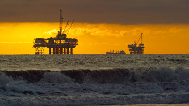 onde che si infrangono sulle coste con diverse sagome di piattaforme di perforazione petrolifera offshore e una petroliera (petroleum) sull'orizzonte in lontananza al tramonto sotto un drammatico, stormy sky - oil rig sea oil storm foto e immagini stock