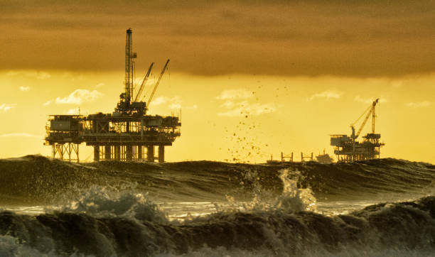waves crashing and splashing near huntington beach in southern california with several silhouettes of offshore oil drilling rig platforms and an oil (petroleum) tanker on the horizon in the distance at sunset under a dramatic, stormy sky - oil rig sea oil storm stock-fotos und bilder
