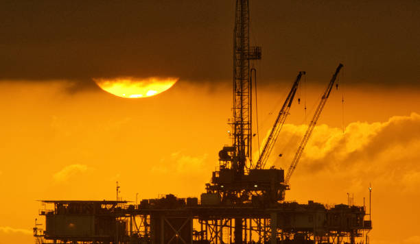 die sonne blickt unter sturmwolken mit einer silhouette einer offshore-ölbohrplattform im vordergrund bei sonnenuntergang unter einem dramatischen, stürmischen himmel hervor - oil rig sea oil storm stock-fotos und bilder