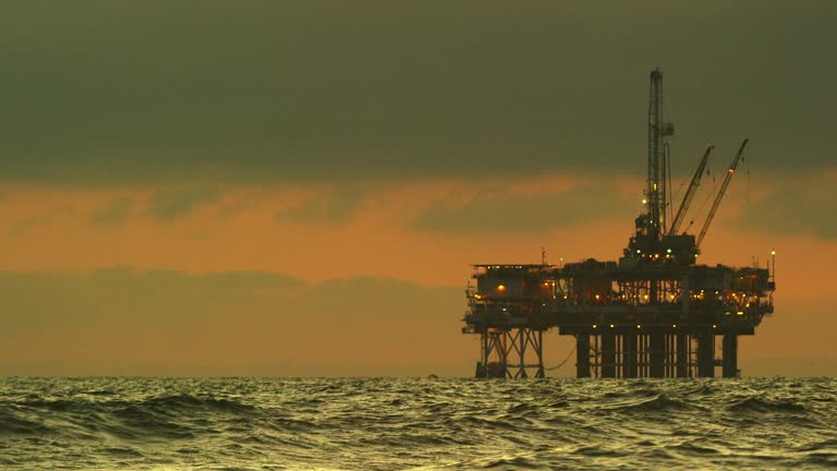 Waves Crash on the Shore of Huntington Beach in Southern California with an Offshore Oil Drilling Rig Platform on the Horizon at Sunset under a Dramatic, Stormy Sky