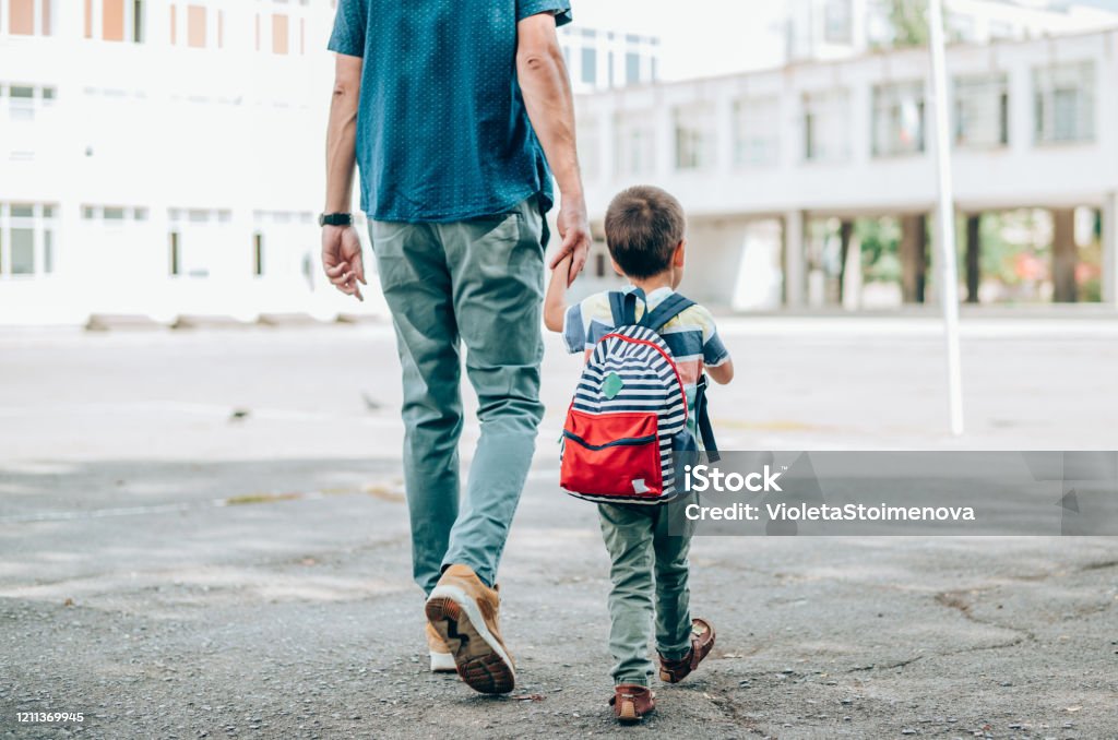 Father and son going to school. Rear view of father who leads a little boy hand in hand to the school. Father and son with backpack walking in schoolyard. Child Stock Photo