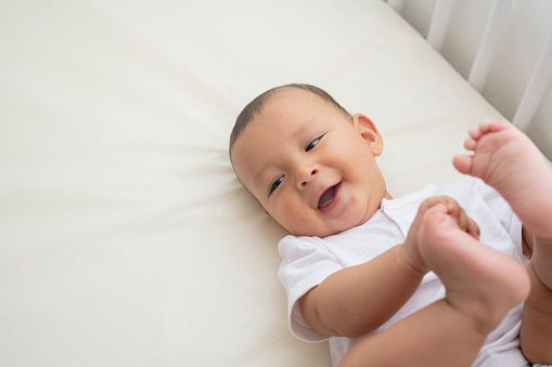 6 month old Latino baby boy dressed in white body lying in his white crib looks sideways shows a beautiful smile as he grabs his feet with his hands