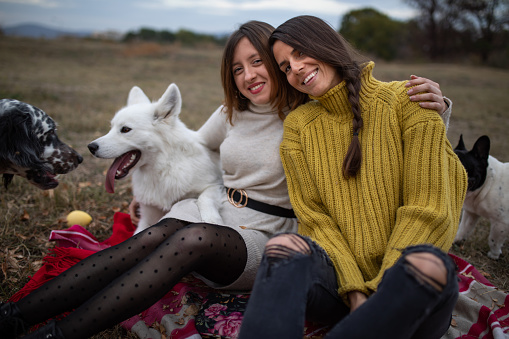 Two girls are sitting on a blanket on a field smiling surrounded by their playful dogs