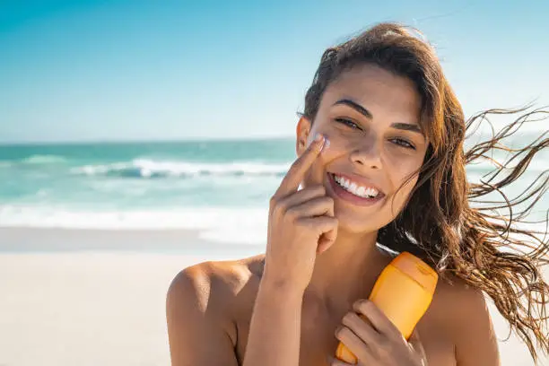 Beautiful young woman at beach applying sunscreen on face and looking at camera. Beauty latin girl enjoying summer holiday while applying suntan lotion at sea. Portrait of happy woman with healthy skin applying sunblock on cheek.