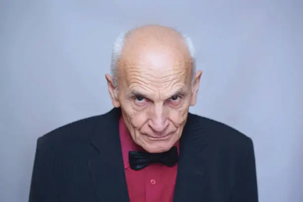 senior elderly man in a black suit and a red shirt with a bow tie stands with his head bowed, looking out from under his forehead, entertainer, singer or actor, a leading party entertainment event