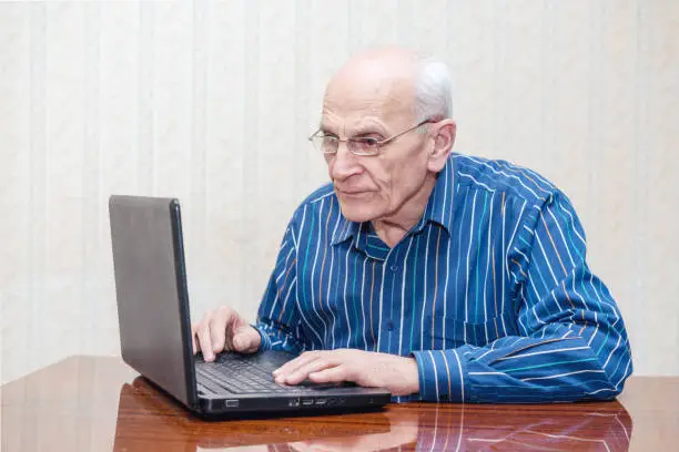elderly man with glasses sits at a table and looks into a laptop
