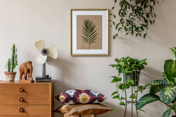 Stylish and vintage interior design of living room with retro furnitures and elegant personal accessories. Mock up poster frame. Template. Home decor. elegante puesta en escena retro de la sala de estar con marco de póster de maqueta de oro, muebles de diseño, una gran cantidad de plantas, almohadas, ventilador retro y accesorios personales elegantes. decoración de casa vintage. plantilla.