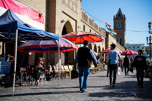 Iraq, Iraqi Kurdistan, Arbil, Erbil. Next to the park Shar men are walking down an alley next to the archs of the entrance of Erbil's bazar.  On the left, a woman is sitting having tea. In the background, stands Erbil's clock.