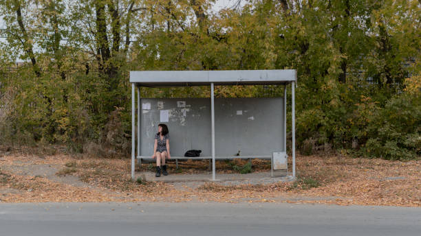 una chica solitaria con un vestido y botas se sienta en una parada de autobús - estación de autobús fotografías e imágenes de stock