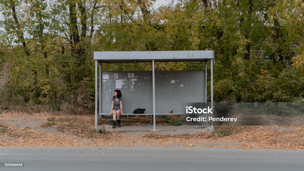 A lonely girl in a dress and boots sits at a bus stop A lonely girl in a dress and boots sits at a bus stop. Bus Stop Stock Photo