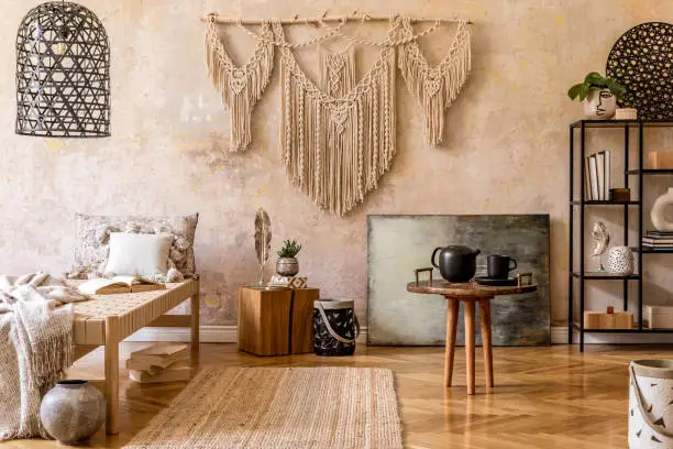 Photo of Stylish oriental living room with design beige chaise longue, beautiful macrame, pouf, pillows, decor, shelf with elegant personal accessories, pendant lamp. Wabi sabi concept. Grunge wall. Home decor