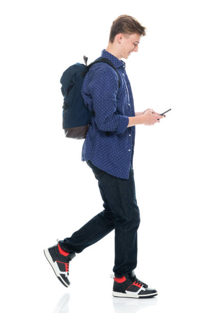 istock Boys student walking in front of white background wearing backpack and holding textbook and using mobile phone 1211431549