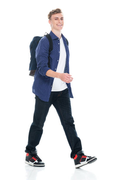 Side view of aged 16-17 years old with brown hair caucasian boys university student walking in front of white background wearing backpack who is laughing and holding bag