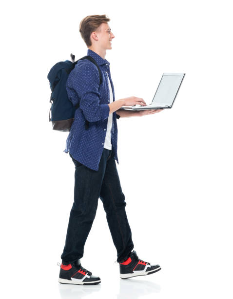 istock Boys student walking in front of white background wearing backpack and holding textbook and using laptop 1211433233