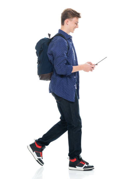 istock Boys student walking in front of white background wearing backpack and holding textbook and using digital tablet 1211433263