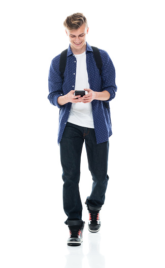 One person of aged 16-17 years old with brown hair boys student walking in front of white background wearing backpack who is learning and holding textbook and using mobile phone