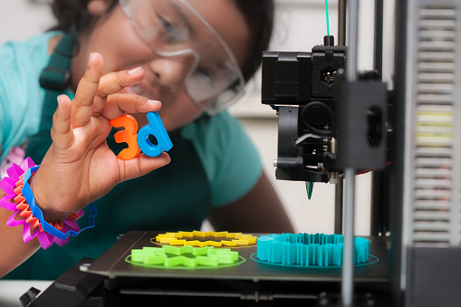 Una estudiante latina inteligente con formas impresas en 3D multicolores como joyas, junto a una impresora 3D con diseños en la cama de impresión calentada, y sosteniendo letras 3d en su mano. photo