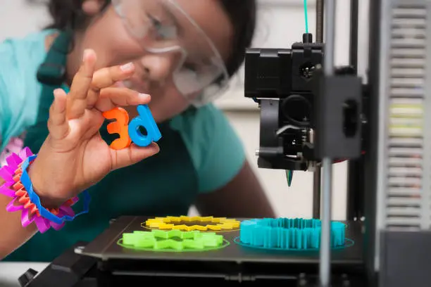 Photo of A smart latino student wearing multi-colored 3d printed shapes as jewelry, next to a 3d printer with designs on the heated print bed, and holding 3d letters in her hand.