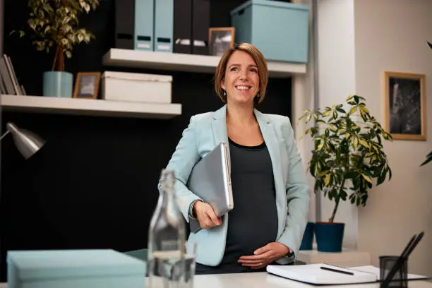 Cute caucasian pregnant businesswoman standing in her office, holding notebook and preparing to go on meeting with her colleagues.