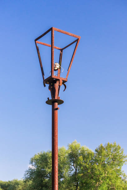 Old rusty street lamp with broken bulb and torn electrical wires against bright, summer blue sky and green trees. Devastation and abandonment of village infrastructure.