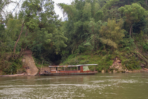 Guilin, China - May 10, 2010: Along Li River. Ramshackled and rusty ferry anchored near shoreline covered with dense forest. Brown path reaches water.