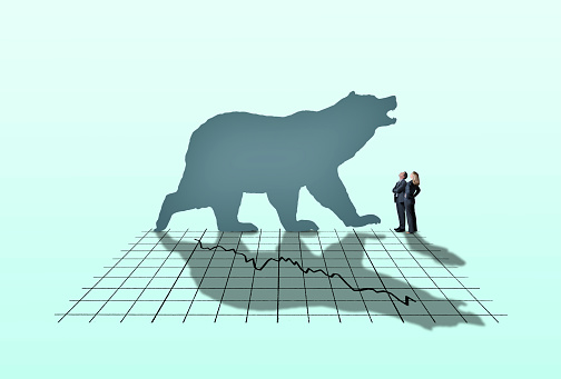 A businessman and a businesswoman look up at a large bear as it casts its shadow across a stock market chart.