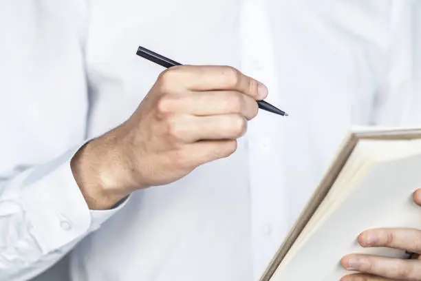 Photo of Man writes standing with a pen in notebook in a sunny office, business and education concept. Close up