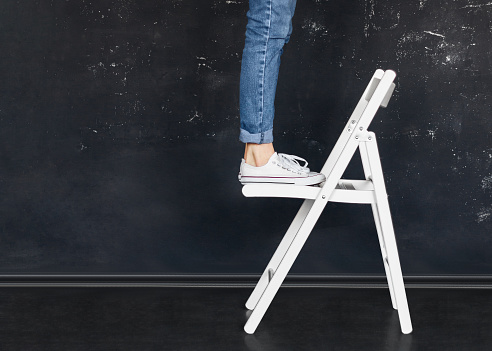 Woman in jeans and white sneakers is standing on a white folding chair in a black room