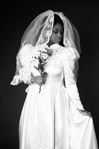 Vertical B&W studio shot on dark background of young woman in 80s style wedding dress, with bouquet and veil. Standing front view with face turned profile, holding out skirt.