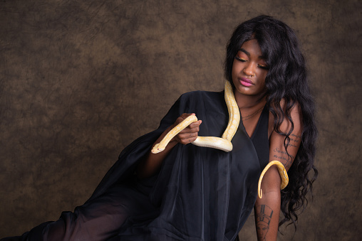 Horizontal studio shot on brown of young woman with long black wavy hair holding snake.