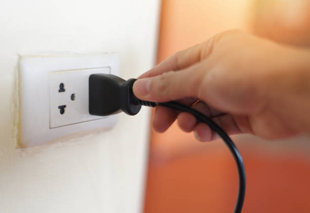 istock Plug in wall with hand and black power cord cable - Unplug or plugged in concept socket on electric plug on wall 1211466443