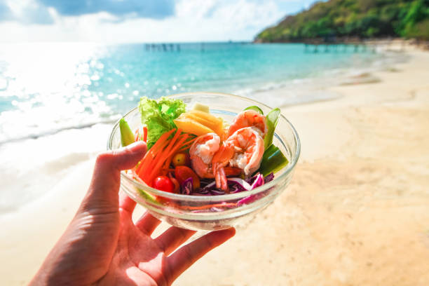 insalata vegetale a portata di mano e sfondo costa del mare - insalata gamberetti con frutta e lattuga fresca pomodoro peperone di carota sulla ciotola cibo sano sulla spiaggia per concetto di festa - pepper bell pepper portion vegetable foto e immagini stock