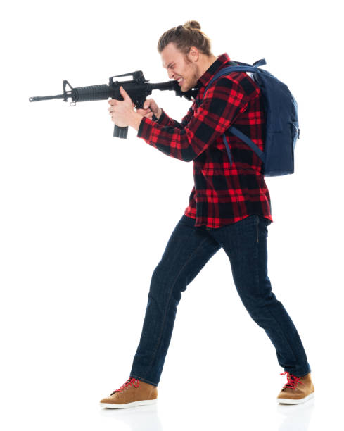 istock Caucasian young male student in front of white background wearing backpack and holding weapon and using machine gun 1211480430